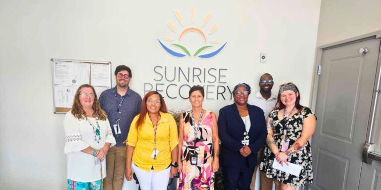 A smiling clinical team of seven professionals (five women and two men) stands in front of a sign that reads "SUNRISE RECOVERY," providing addiction treatment services. The team is diverse and professionally dressed, positioned beneath the company's logo.