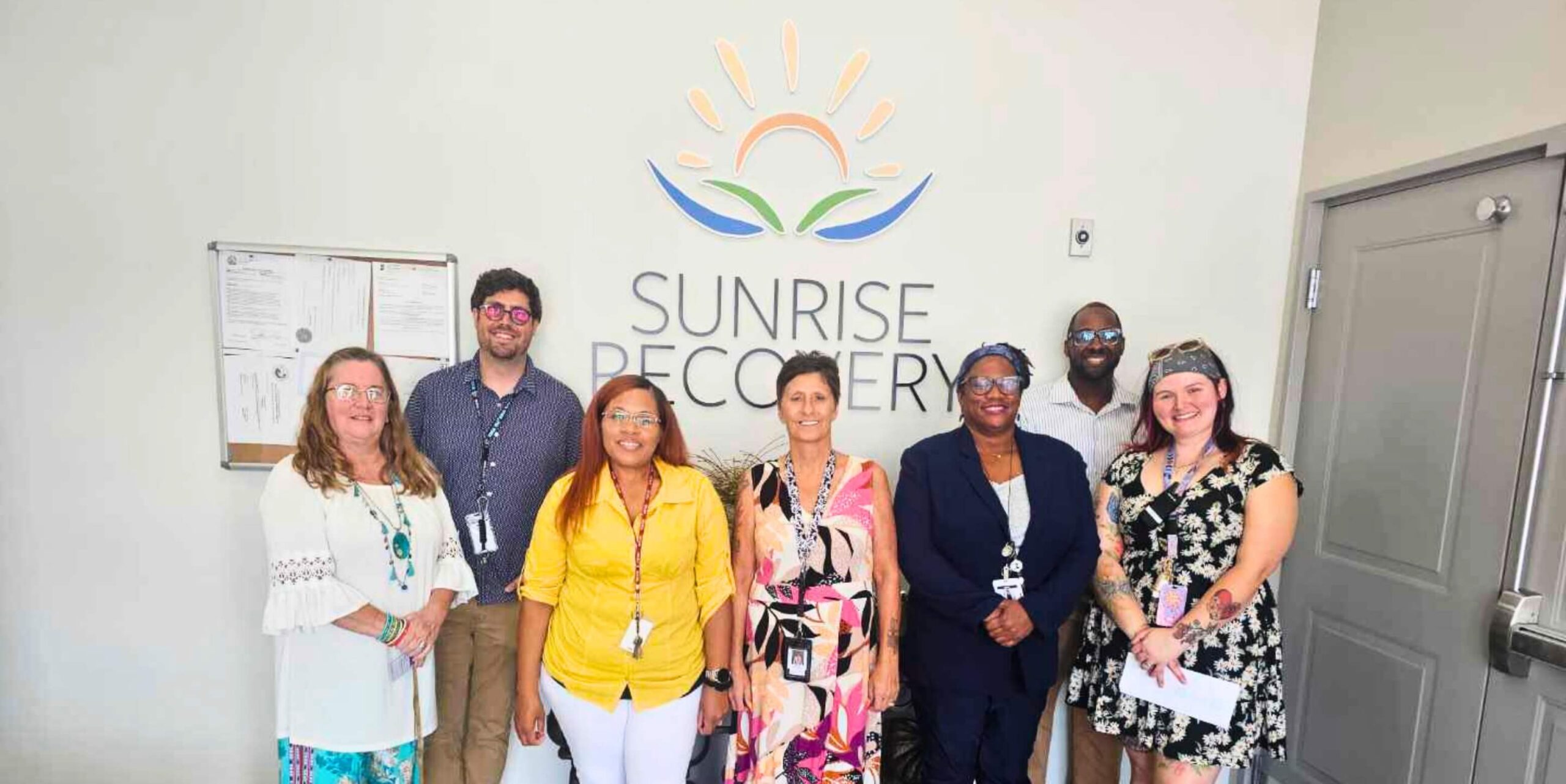 A smiling clinical team of seven professionals (five women and two men) stands in front of a sign that reads "SUNRISE RECOVERY," providing addiction treatment services. The team is diverse and professionally dressed, positioned beneath the company's logo.