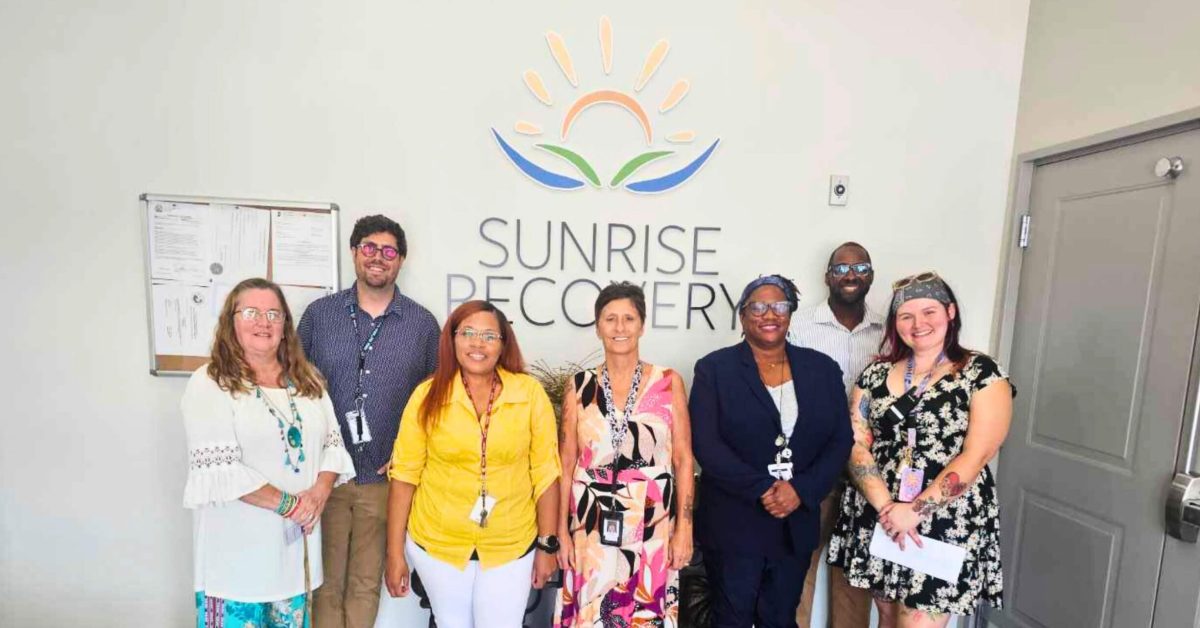 A smiling clinical team of seven professionals (five women and two men) stands in front of a sign that reads "SUNRISE RECOVERY," providing addiction treatment services. The team is diverse and professionally dressed, positioned beneath the company's logo.